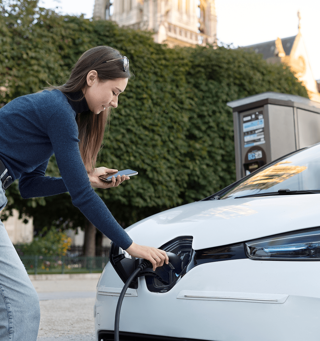 A young woman bending over electric vehicle, looking at a smartphone in her hands, possibly using a mobile app to initiate the charging process.