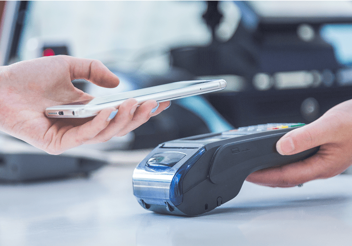A person's hand holds a smartphone above acredit card terminal for contactless payment.